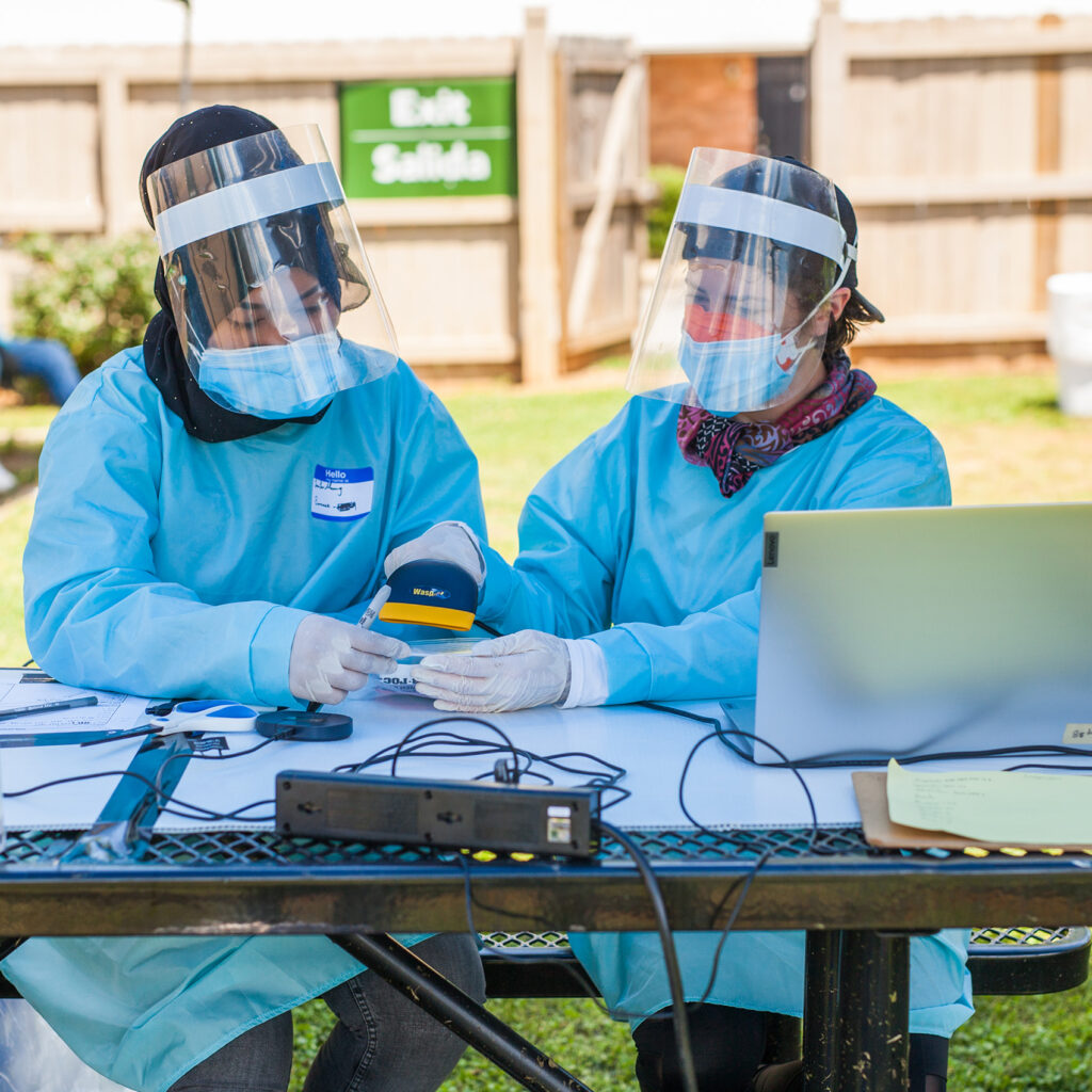 Two CORE staff dressed in full PPE register a COVID test at the table.