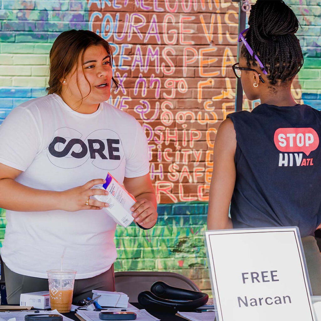 A CORE staff member at a CORE booth talking to someone about Narcan. A sign sits on the table saying "Free Narcan."