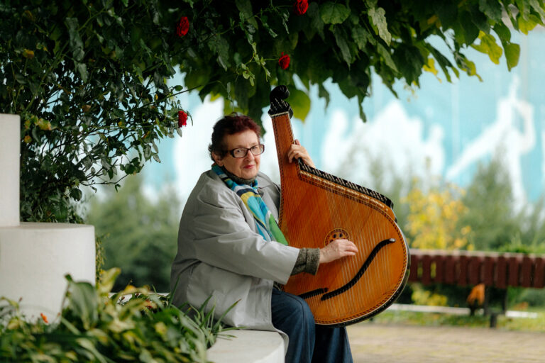 Music teacher, Halyna, sits outside on a bench, under a tree holding a traditional Ukrainian instrument known as a bandura.