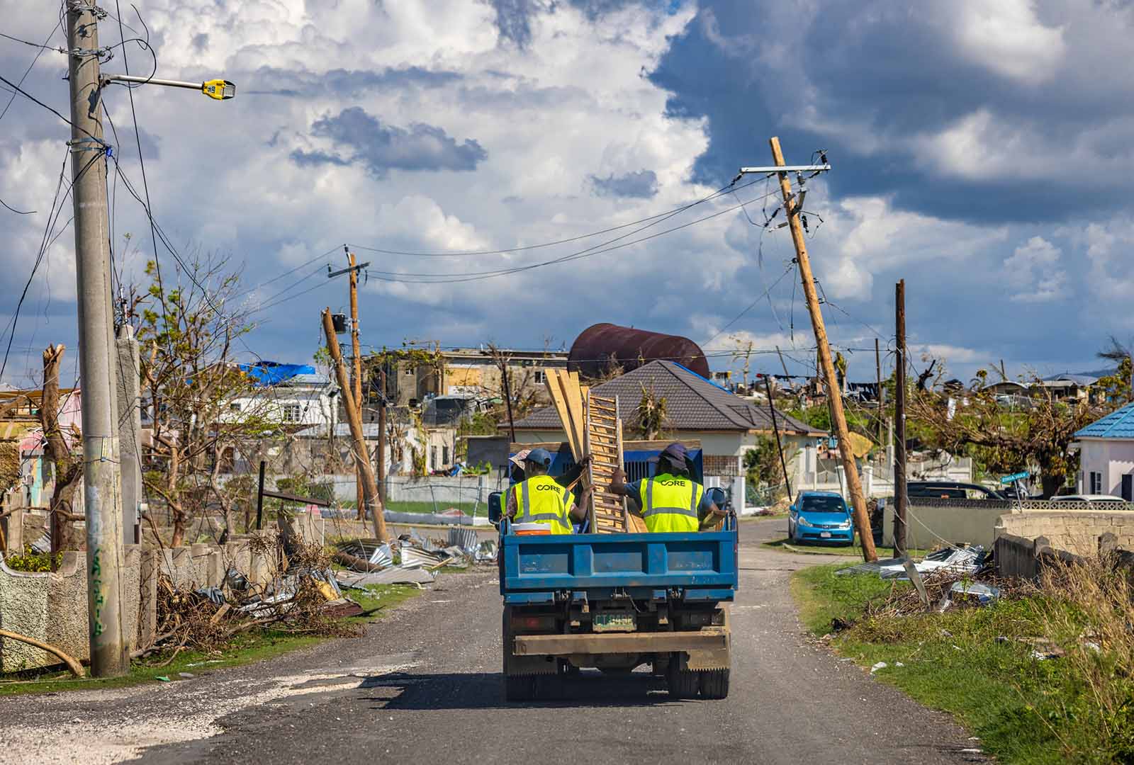 A truck with men sitting in the back drives through a neighborhood damaged by Hurricane Melissa.