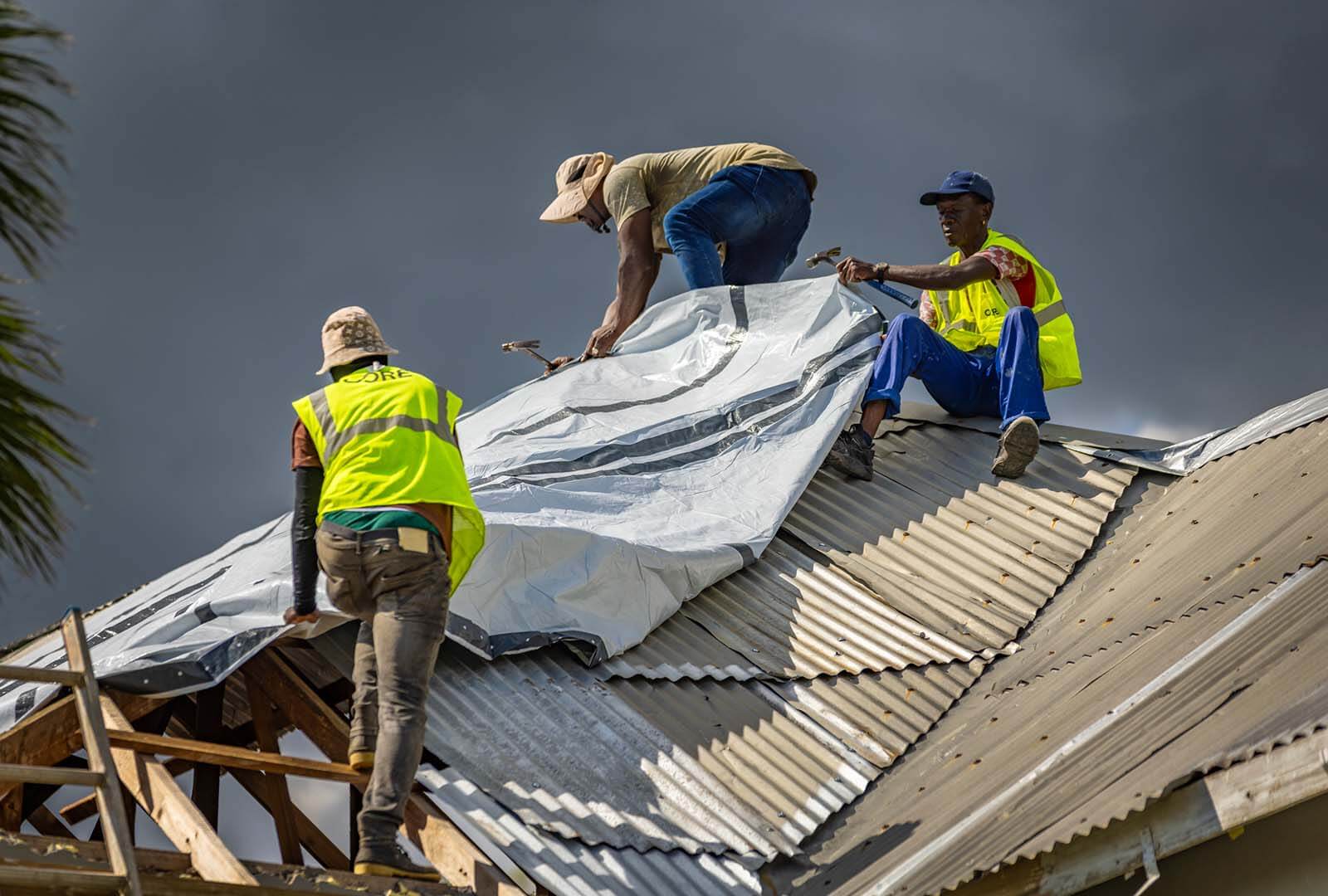 Three men on a mental roof, adding a tarp to prevent rain water from getting in.