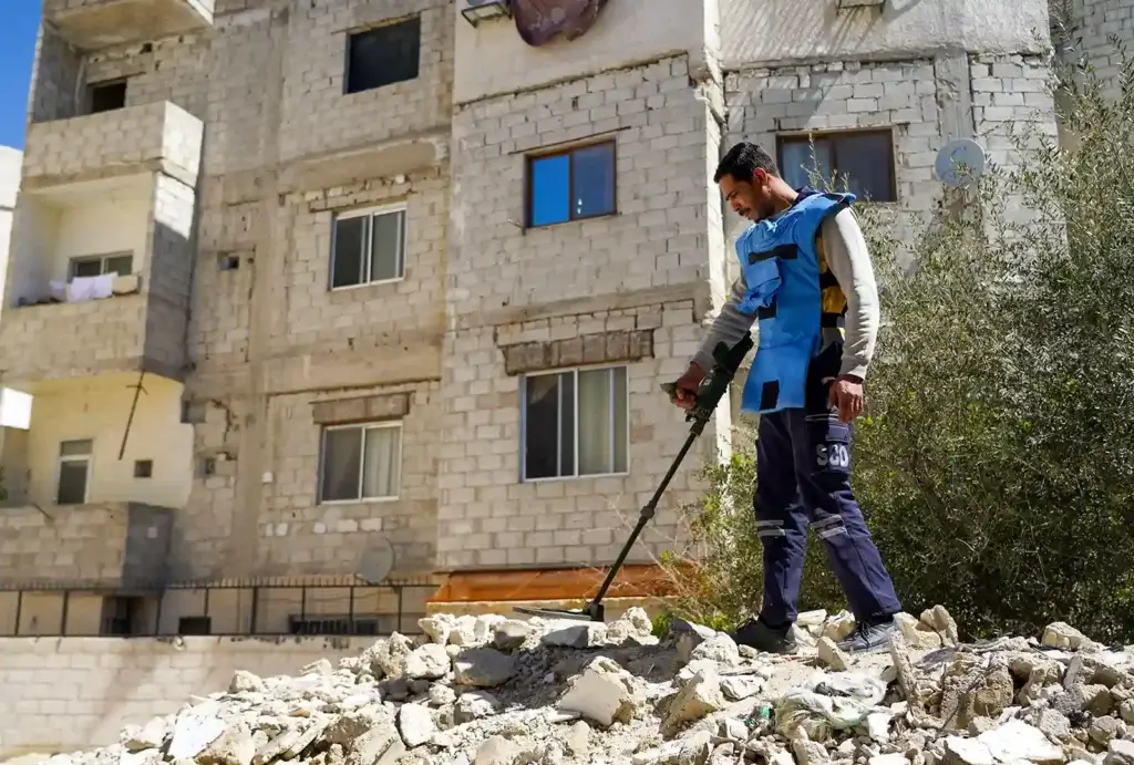 A member of the unexploded ordnance team is wearing a protective gear while holding mental dectors as he walk over rubble.